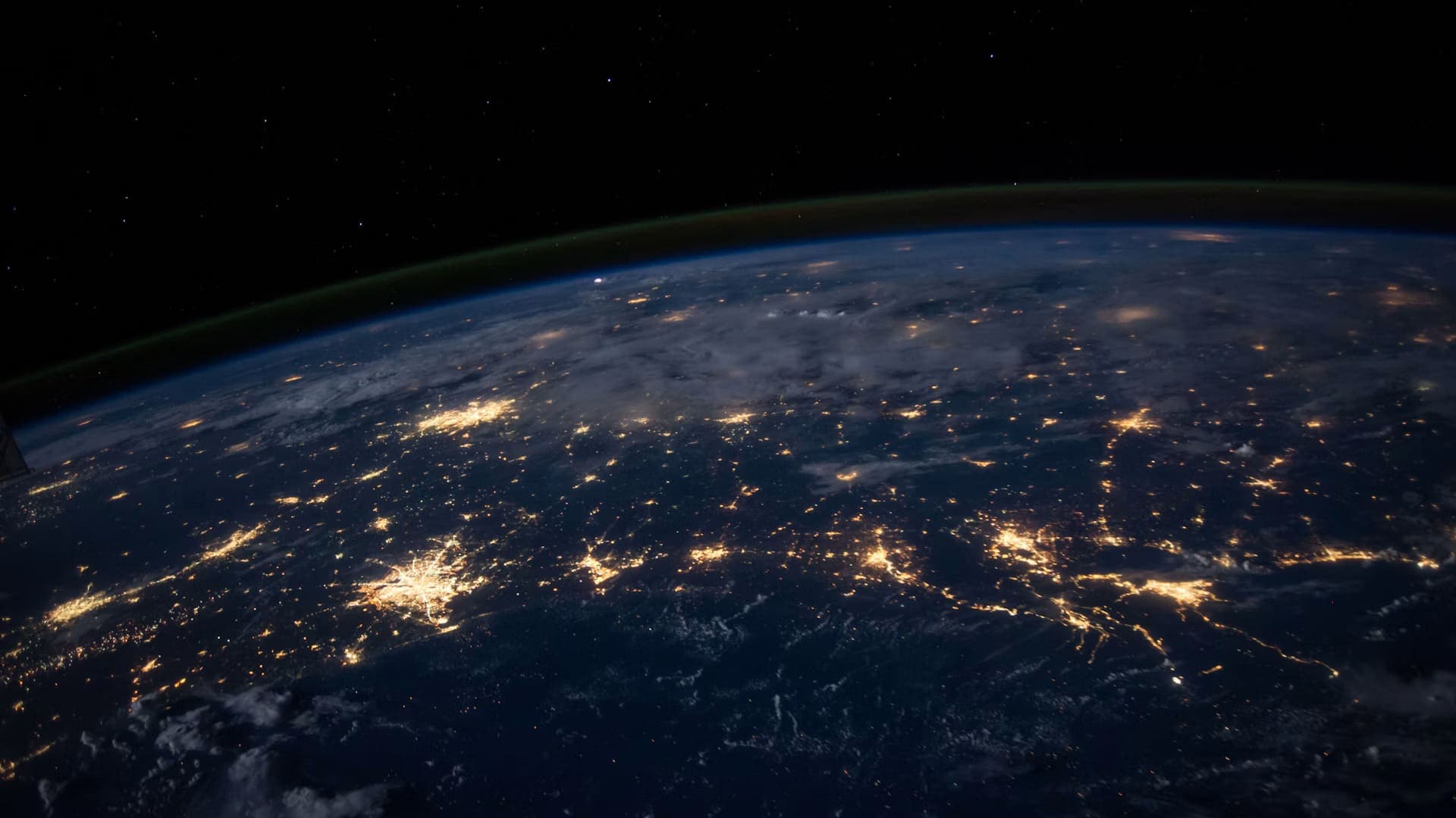 The Earth at night photographed from orbit, city lights spreading in connected networks across the darkened surface beneath a thin band of atmosphere.